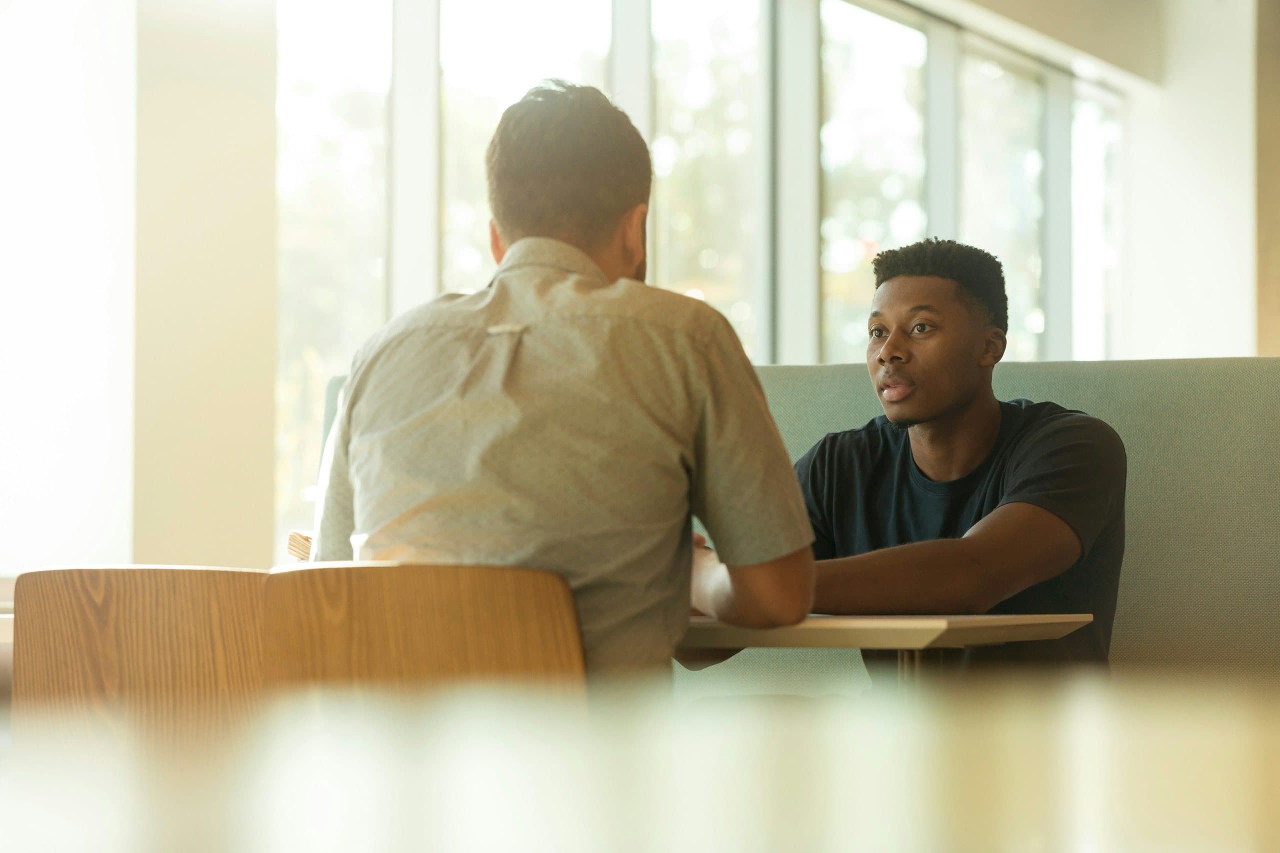 Two men in conversation at a table by a sunlit window, one of them listening