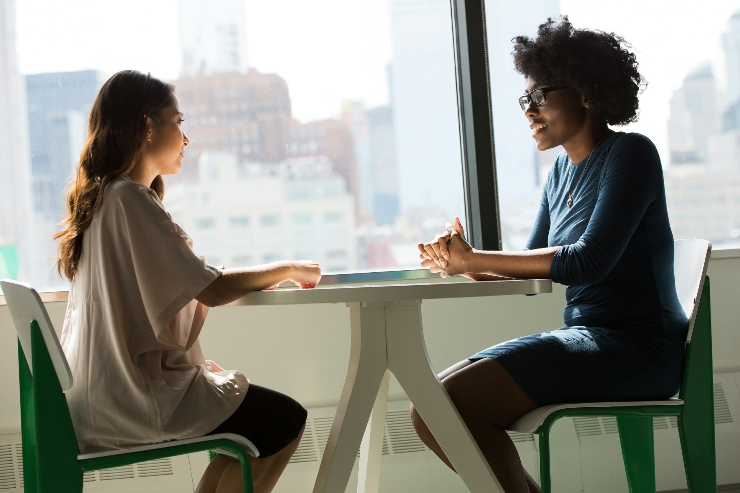 Two women talking across a table by a large window